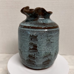 May include: A handmade ceramic vase with a unique ruffled rim. The vase features a blend of blue and brown hues, with a textured surface. The vase is a decorative item, perfect for displaying flowers or as a standalone art piece.