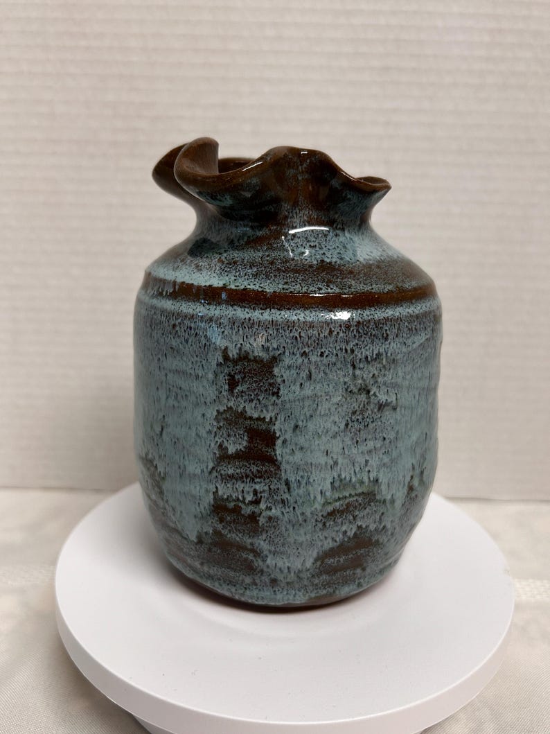 May include: A handmade ceramic vase with a unique ruffled rim. The vase features a blend of blue and brown hues, with a textured surface. The vase is a decorative item, perfect for displaying flowers or as a standalone art piece.