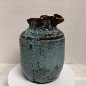 May include: A handmade ceramic vase with a unique, ruffled rim. The vase features a blend of blue and brown glaze, creating a textured, organic appearance. The vase is approximately 5 inches tall.