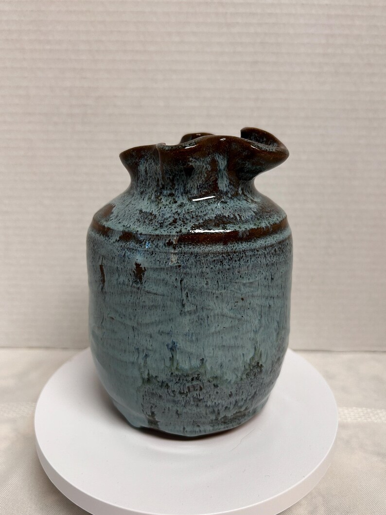 May include: A handmade ceramic vase with a unique, ruffled rim. The vase features a blend of blue and brown glaze, creating a textured, organic appearance. The vase is approximately 5 inches tall.