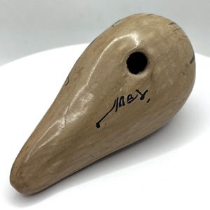 May include: A beige ceramic ocarina with a teardrop shape. It features a single hole and black markings. The ocarina has a smooth, glossy finish and a signature in black ink.
