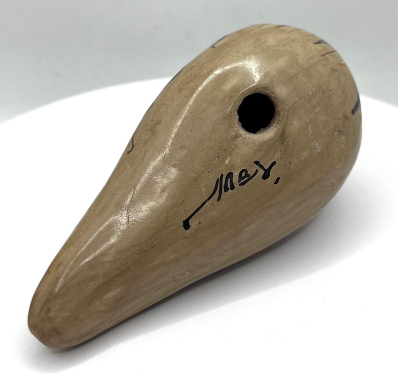 May include: A beige ceramic ocarina with a teardrop shape. It features a single hole and black markings. The ocarina has a smooth, glossy finish and a signature in black ink.