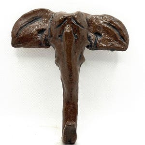 May include: A brown elephant-shaped wall hook. The hook is made of a textured material and features detailed ears, eyes, and a trunk. The hook is designed to be mounted on a wall for hanging items.