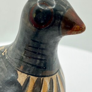 May include: A ceramic bird figurine with a black head and body, featuring a brown beak and red eye details. The bird's neck has black stripes, and the lower body has a pattern of brown and beige vertical lines. The figurine is set against a white background.