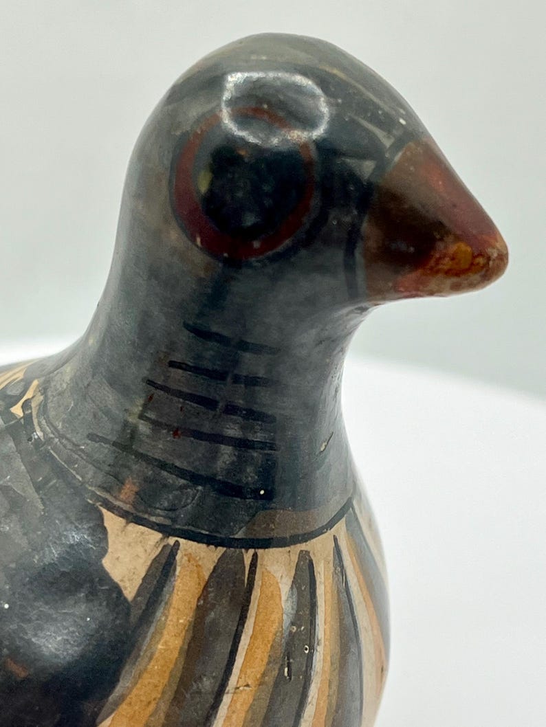 May include: A ceramic bird figurine with a black head and body, featuring a brown beak and red eye details. The bird's neck has black stripes, and the lower body has a pattern of brown and beige vertical lines. The figurine is set against a white background.