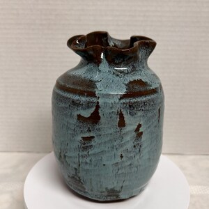 May include: A small, handcrafted ceramic vase with a ruffled rim. The vase features a textured, teal-colored body with brown accents and a dark brown rim. The vase is displayed on a white, circular stand.