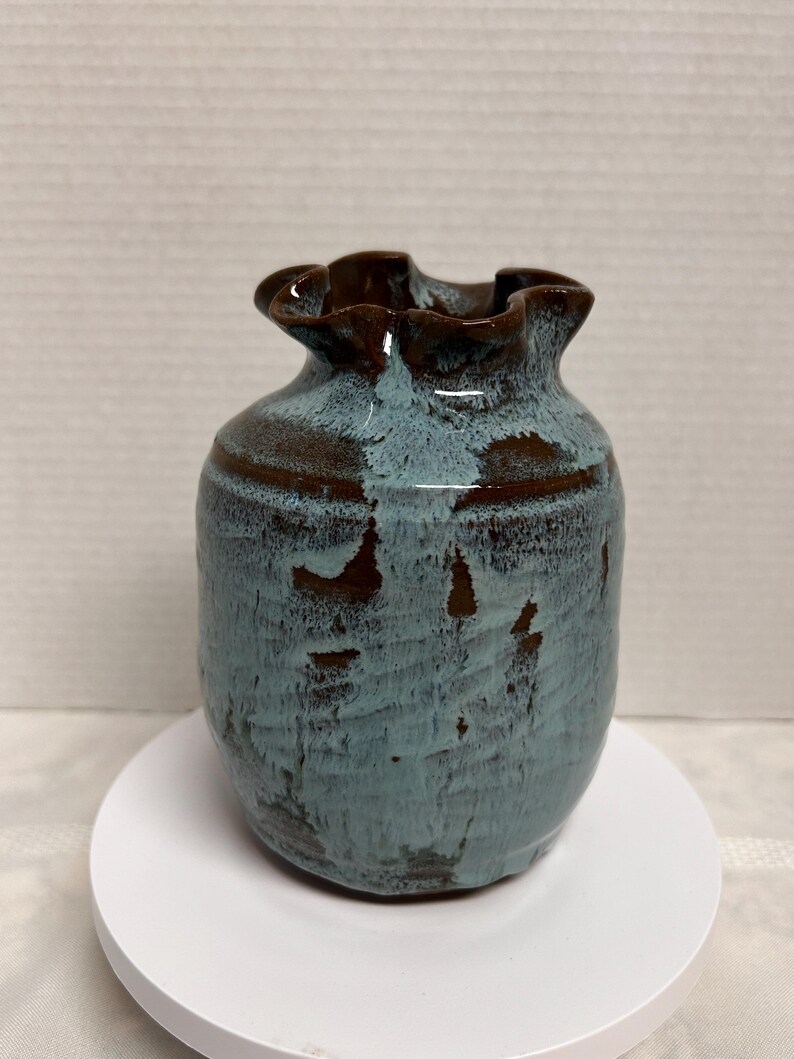 May include: A small, handcrafted ceramic vase with a ruffled rim. The vase features a textured, teal-colored body with brown accents and a dark brown rim. The vase is displayed on a white, circular stand.