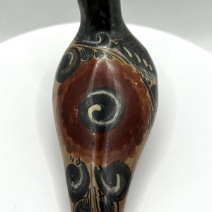 May include: Hand-painted ceramic vessel with a teardrop shape. The top is black, transitioning to a tan body with a central maroon floral design and black and white accents. The vessel is likely for decorative use.