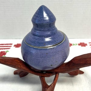 May include: A small, round, blue ceramic jar with a conical lid. The jar sits on a dark brown wooden stand with three branches. The jar has a glossy finish and a light-colored rim.
