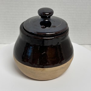 May include: A small, handcrafted ceramic sugar bowl with a lid. The bowl is dark brown with a lighter tan base. The lid has a small, round knob. The sugar bowl is likely used for storing sugar or other dry ingredients.