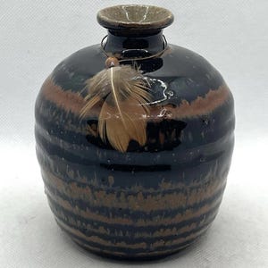 May include: A handcrafted ceramic vase with a dark blue and brown striped pattern. The vase has a small opening and is adorned with a feather and a wire. The vase is approximately 5 inches tall.