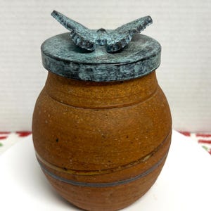 May include: A handcrafted, brown ceramic jar with a lid featuring a butterfly design. The jar has a round shape with a textured surface and a dark blue-green lid. The butterfly is also dark blue-green.