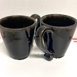 May include: Two dark blue ceramic mugs with a shared handle. The mugs have a glossy finish with a brown rim and small red floral accents. The mugs are handmade and suitable for coffee or tea.