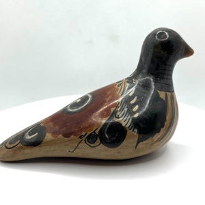 May include: A ceramic bird figurine with a black head and a body featuring brown, tan, and black painted designs. The bird has a long, curved body and a small beak. The figurine is set against a white background.