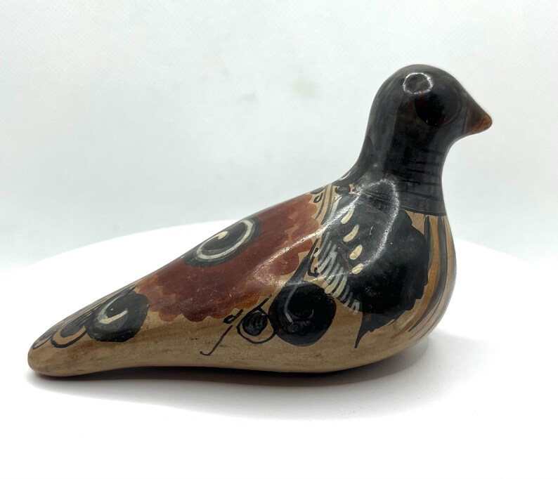 May include: A ceramic bird figurine with a black head and a body featuring brown, tan, and black painted designs. The bird has a long, curved body and a small beak. The figurine is set against a white background.