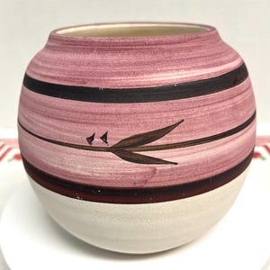 May include: A round ceramic vase with a wide opening. The vase features a white base, a dark brown band, and a pink body with black stripes. A brown leaf design is painted on the pink surface. The vase is likely used for decorative purposes.