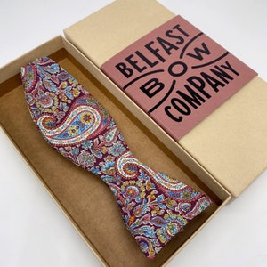 Può includere: Un papillon a fantasia paisley in una scatola. Il papillon presenta uno sfondo bordeaux con disegni floreali blu, gialli e bianchi. La scatola è beige con un'etichetta bordeaux con la scritta "Belfast Bow Company".