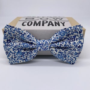 Op de afbeelding: Een witte strik met een blauw bloemenpatroon. De strik ligt op een bruine doos met de tekst "BELT BOW COMPANY" erop gedrukt.