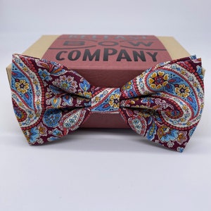 May include: A burgundy bow tie with a paisley pattern in shades of blue, pink, and yellow. The bow tie is on a brown box with the text "Relay Bow Company" printed on it.