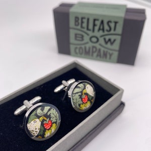 May include: A pair of silver cufflinks with a black background and a colorful bird and berry design. The cufflinks are in a black box with a green label that reads "Belfast Bow Company".
