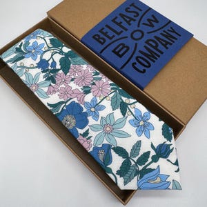 May include: A white tie with a floral pattern in shades of blue, pink, and green. The tie is in a brown box with a blue label that reads "Belfast Bow Company".