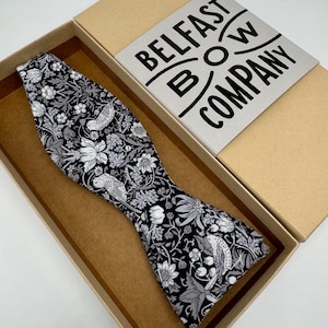 May include: A black bow tie with a white and gray floral pattern. The bow tie is in a brown box with the text "BELFAST BOW COMPANY" on the lid.