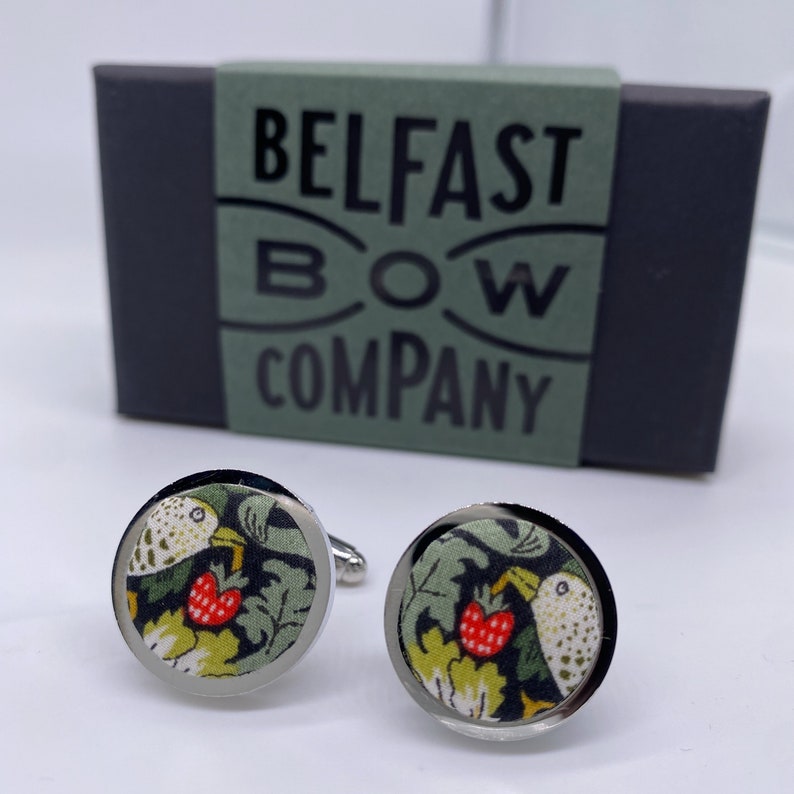May include: A pair of silver cufflinks with a round, black and green fabric design featuring a bird and a red strawberry. The cufflinks are in a box that says "Belfast Bow Company".