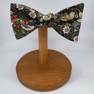 May include: A black bow tie with a green, white, and red floral pattern featuring birds. The bow tie is displayed on a wooden stand.