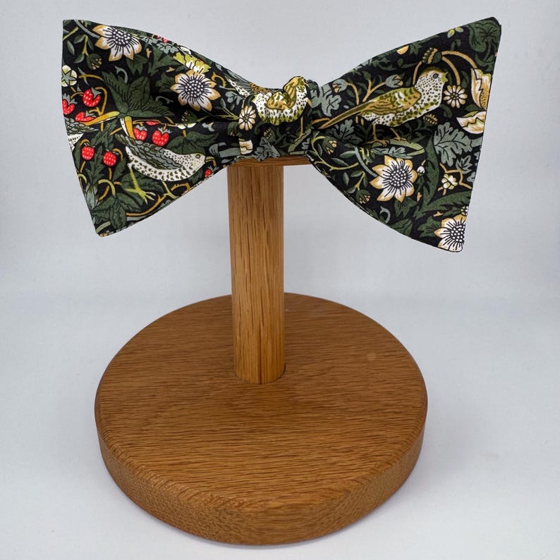 May include: A black bow tie with a green, white, and red floral pattern featuring birds. The bow tie is displayed on a wooden stand.