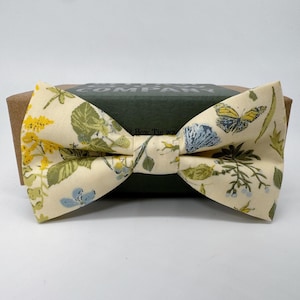 Puede incluir: Una pajarita de color crema con un estampado floral con flores amarillas, verdes y azules y mariposas. La pajarita está sobre una caja marrón con el texto "Bow Tie Company" impreso en ella.