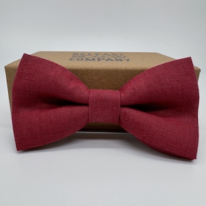 Könnte beinhalten: Ein burgunderfarbener Fliege aus Leinen. Die Fliege liegt auf einem braunen Karton mit dem aufgedruckten Text "Belfast Bow Tie Company".