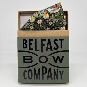 May include: A green box with the text "BELFAST BOW COMPANY" printed on the front. The box contains a green and brown patterned pocket square with a bird and floral design.
