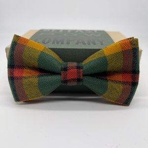 May include: A colourful plaid bow tie in green, yellow, orange, and black. The bow tie sits on a green box with the text "BELFAST BOW COMPANY". The bow tie is a classic shape and made of woven fabric.