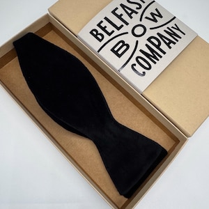 May include: A black velvet bow tie rests inside a cardboard box. The box lid displays the text "Belfast Bow Company" in a circular design. The bow tie is a classic shape, with a soft, textured appearance.