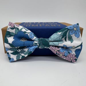 May include: A white bow tie with a floral pattern in shades of blue, pink, and green. The bow tie is on a blue box with the text "Belfast Bow Company" printed on it.