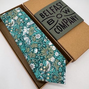 May include: A teal necktie with a floral and bird design, displayed in a brown box. The tie showcases white flowers and birds on a teal and green background. The box has a label that reads "Belfast Bow Company."