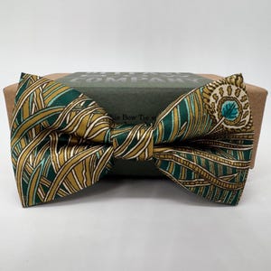May include: A green and gold patterned bow tie with a peacock feather design. The bow tie is on top of a brown box. The bow tie has a repeating pattern of gold and green lines and a circular design on the right side.