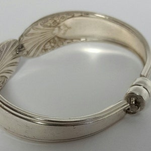 May include: A silver spoon bracelet with an ornate design. The bracelet is made from a bent spoon and has a magnetic clasp.