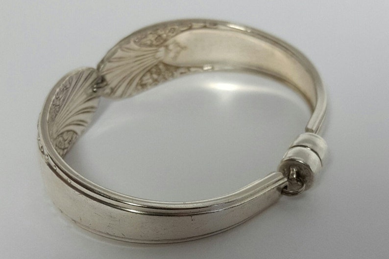 May include: A silver spoon bracelet with an ornate design. The bracelet is made from a bent spoon and has a magnetic clasp.