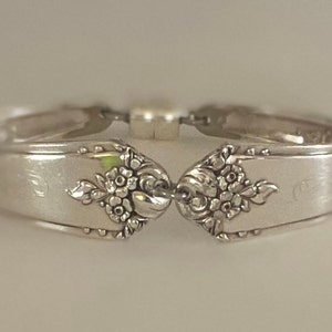 May include: A silver spoon bracelet with floral designs on the ends. The bracelet is made from two spoons that are joined together with a hinge.