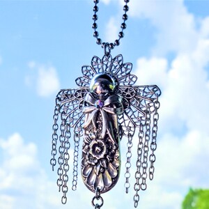 May include: A silver necklace with a detailed angel pendant featuring a filigree design, a bow, and a small iridescent gemstone. The pendant hangs from a chain with multiple dangling chains.