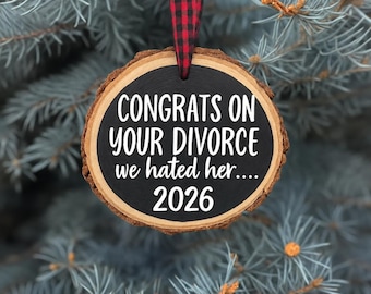 Puede incluir: Un adorno de rodaja de madera redonda con bordes de corteza natural, con un centro pintado de negro y texto blanco que dice: CONGRATS ON YOUR DIVORCE we hated her.... 2026. Cuelga de una cinta de cuadros rojos y negros.