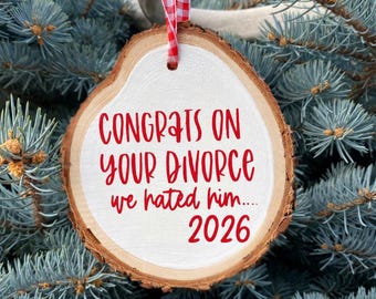 Puede incluir: Un adorno de rodaja de madera redonda con bordes de corteza natural, con un frente pintado de blanco y texto rojo que dice: CONGRATS ON YOUR DIVORCE we hated him.... 2026. Una cinta a cuadros roja y blanca está unida a la parte superior.