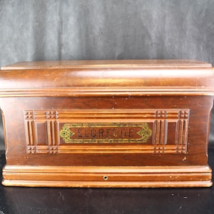 May include: A vintage wooden box with a curved top and decorative panels. The box is a rich brown color and features the name "ELDREDGE" in gold lettering. The box has a keyhole and is likely an antique sewing machine cabinet.