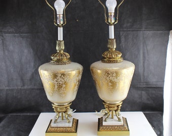 May include: Pair of vintage table lamps with a cream-colored, speckled glass base and gold floral design. The lamps have brass accents, including the base, decorative crown, and lamp holder. Each lamp has a white bulb and a square marble base.