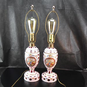 May include: A pair of table lamps featuring white and cranberry red cut-to-clear glass bases with hand-painted oval medallions depicting figures in period clothing. Each lamp has a brass harp, a brass socket, and an exposed Edison-style bulb.
