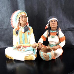 May include: Two Native American figurines. One figure wears a feathered headdress and a patterned tunic. The other figure wears a headband and holds a woven basket. Both figures are seated and have detailed facial features.