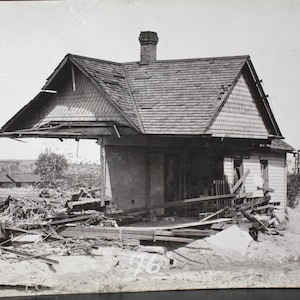 May include: A black and white photograph of a damaged house. The roof is partially collapsed, and debris litters the ground. The house has a chimney and a small porch. The number "76" is visible on the ground.