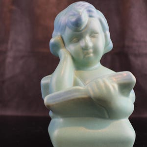 Puede incluir: Una figurita de cerámica azul de un niño leyendo un libro. La figurita está en posición sentada con la cabeza del niño apoyada en la mano.