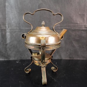 May include: A vintage copper tea kettle with a rounded body, a short spout, and a decorative arched handle. The kettle rests on a tripod stand featuring a small integrated burner at the base, with floral accents on the metal legs and a dark patina.